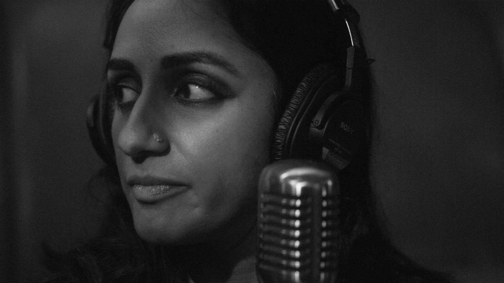 A person wearing headphones looks to the side in front of a vintage-style microphone in a recording studio. The image is in black and white.