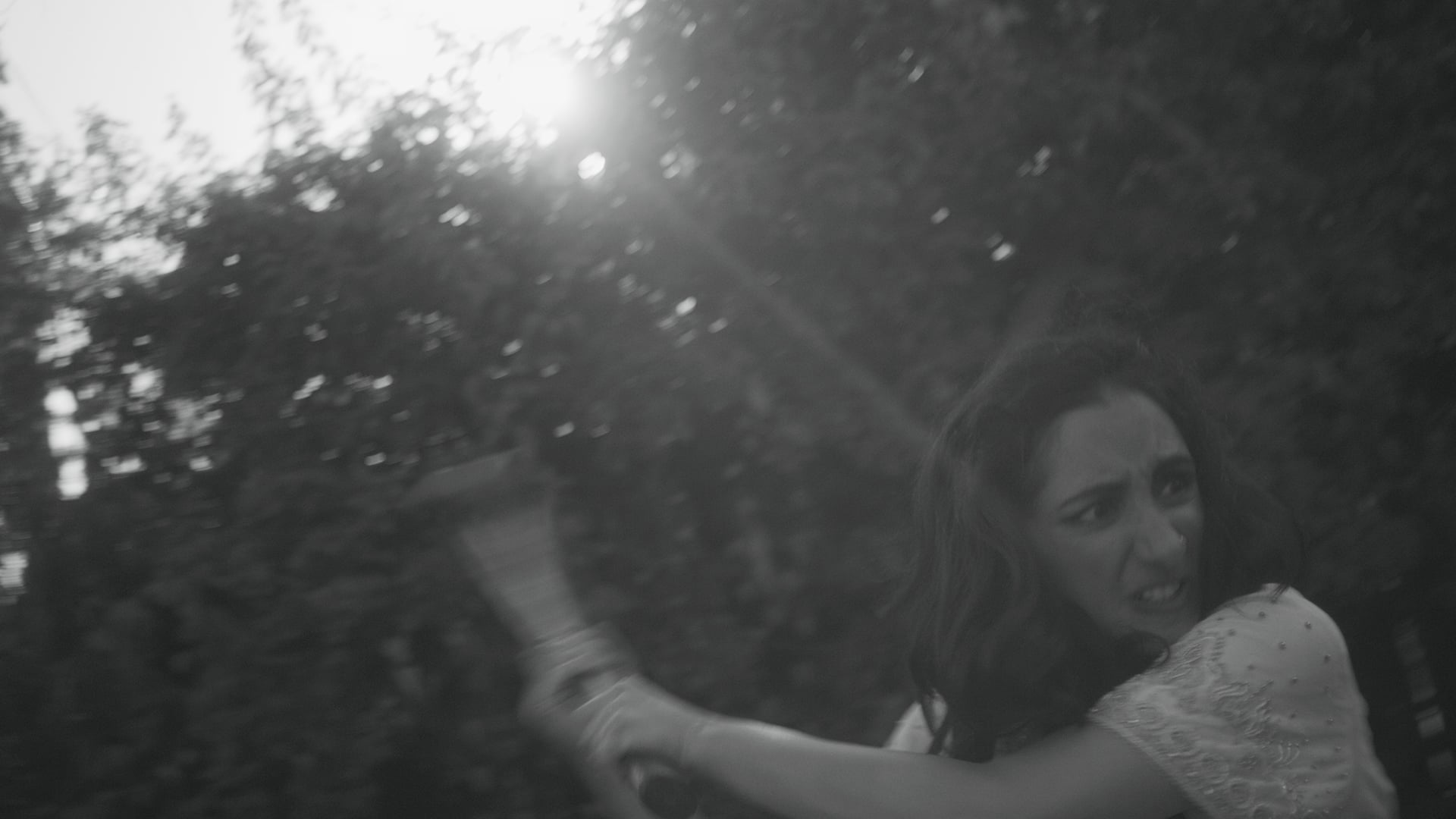 A woman with a tense expression swings an object outdoors, with sunlight and trees in the background. The image is in black and white.