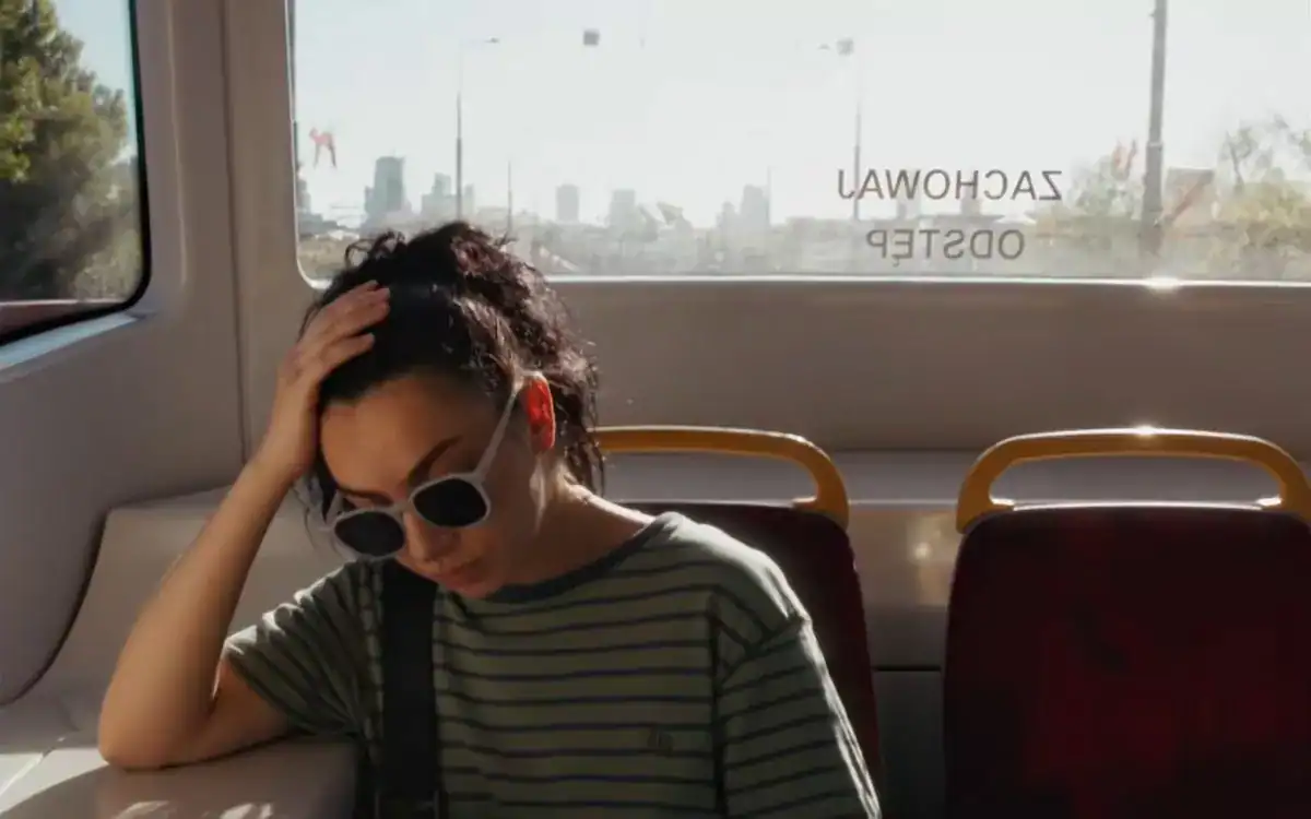 A person wearing sunglasses and a striped shirt sits alone on a bus, resting their head on their hand, with a cityscape visible through the window behind them.