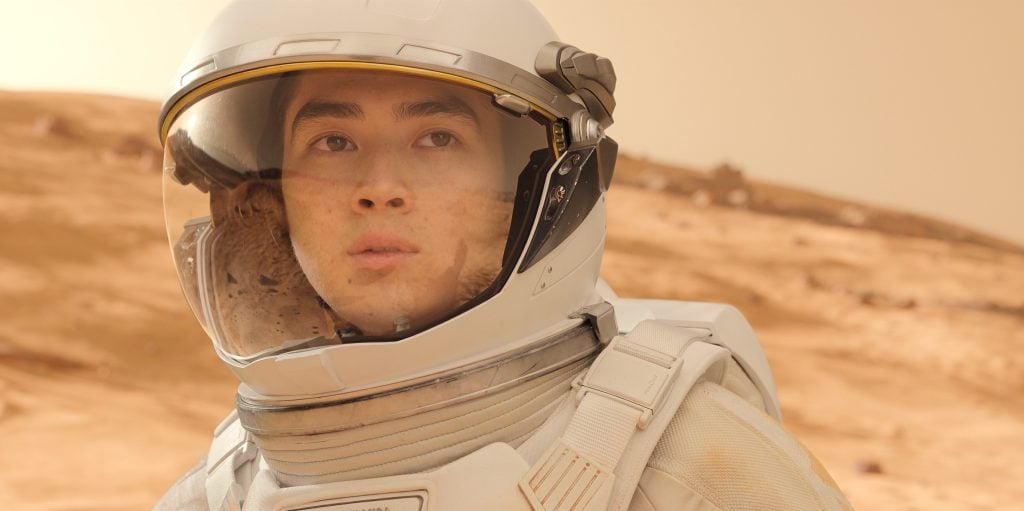 A person wearing a white space suit and helmet stands on a rocky, desert-like terrain under a yellowish sky.