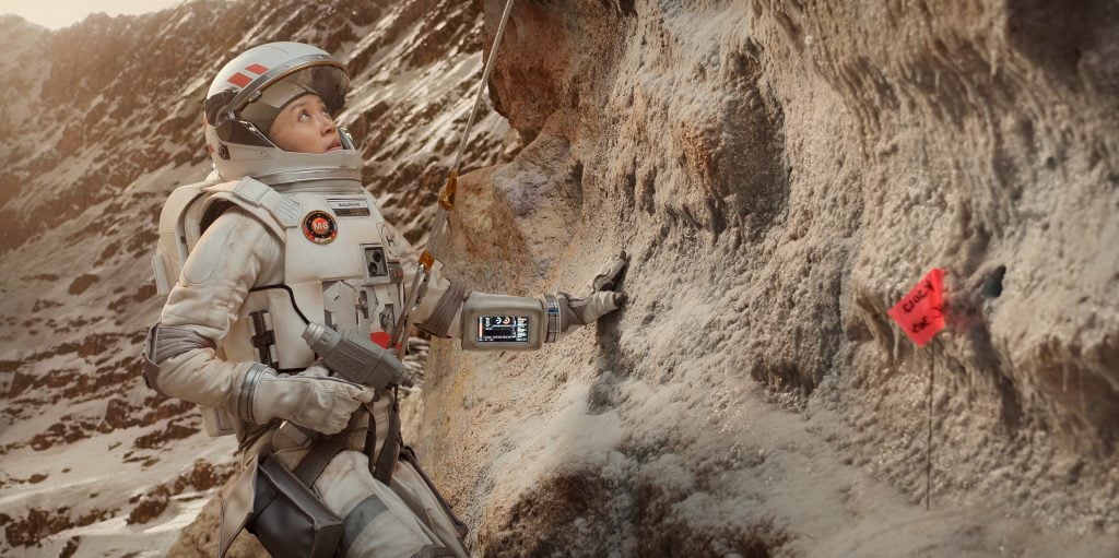 An astronaut in a white space suit examines a rocky, mountainous surface, holding onto the rock near a red flag marker labeled "EXIT.
