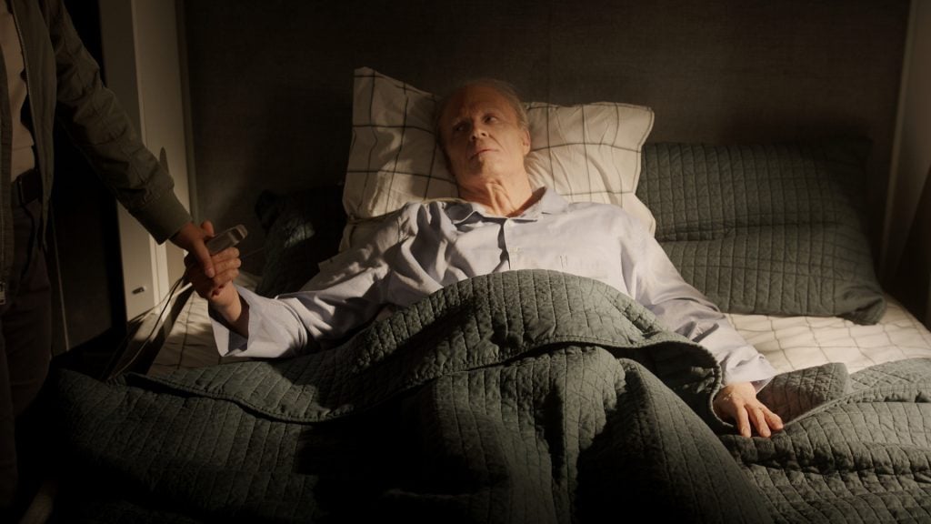 An elderly man lies in bed under a quilt, holding hands with someone standing beside him, looking up with a neutral expression.