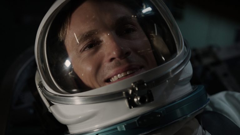 A close-up of a person smiling inside a spacesuit helmet, with reflections visible on the visor.