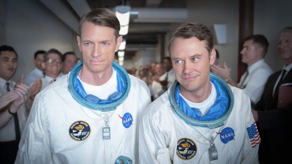 Two astronauts in white NASA suits walk down a hallway as people in the background applaud.