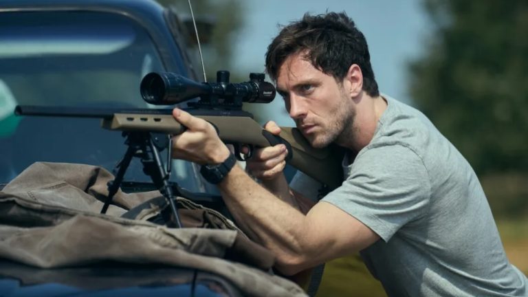 A man in a gray t-shirt aims a scoped rifle using a bipod, resting it on a car next to him, with focused expression outdoors.