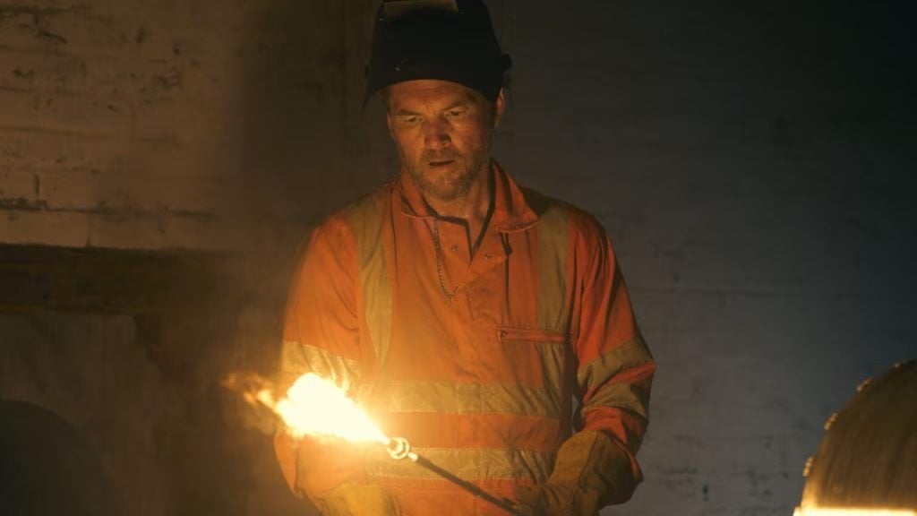 A worker in an orange safety jacket and helmet uses a torch that emits a visible flame in an industrial setting.