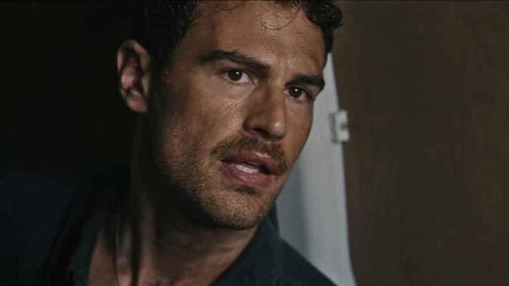 A man with short dark hair and light facial stubble looks intently to the side, partially illuminated, against a plain indoor background.