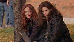 Two young women with serious expressions sit outside on the grass near a tree and brick wall, one holding a laptop.