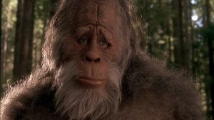 A close-up of a Bigfoot or sasquatch creature with a sad expression, standing in a forest with trees in the background.