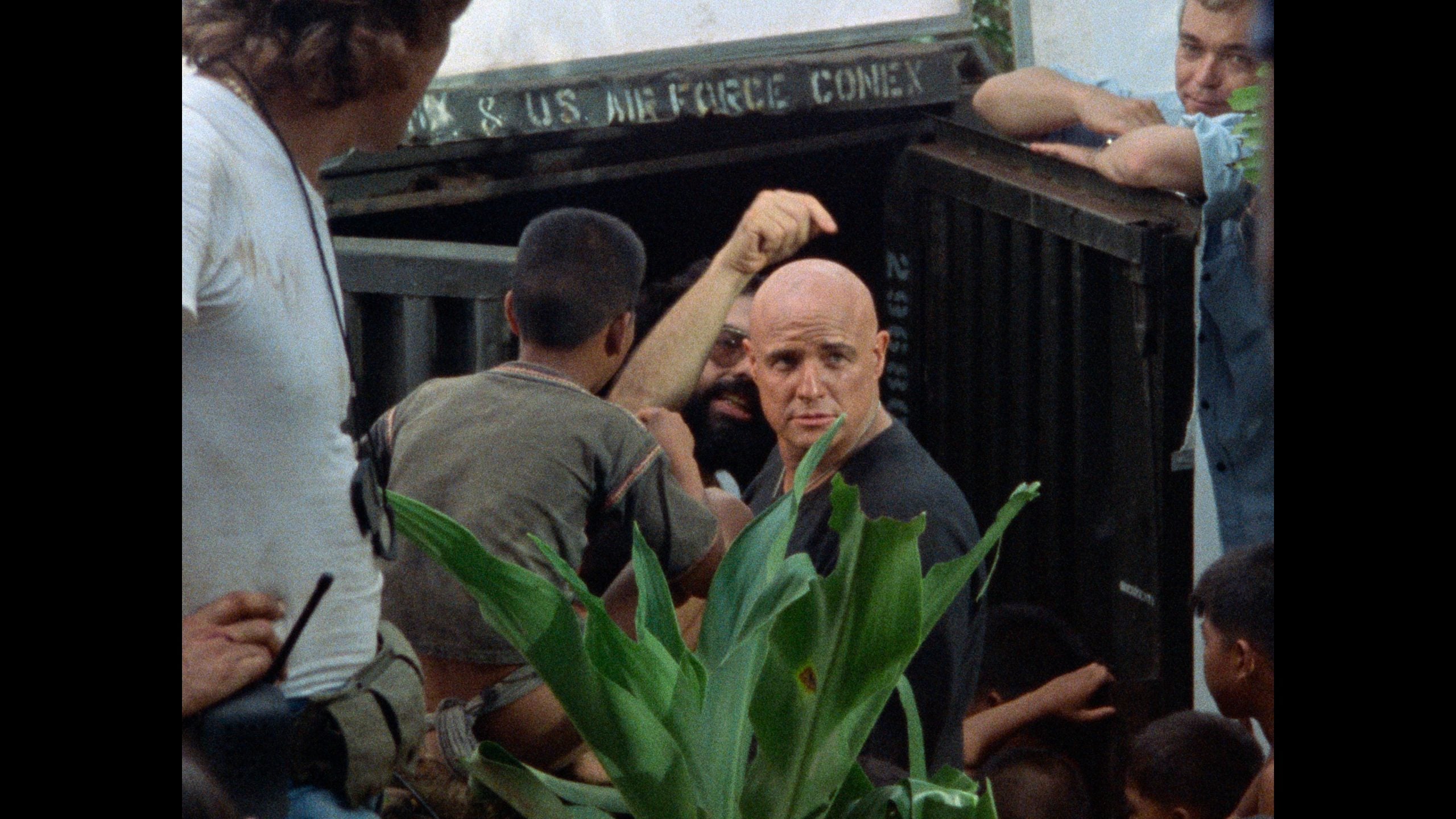 A bald man looks toward the camera while others gather around a container labeled "U.S. Air Force Conex;" green leaves are visible in the foreground.