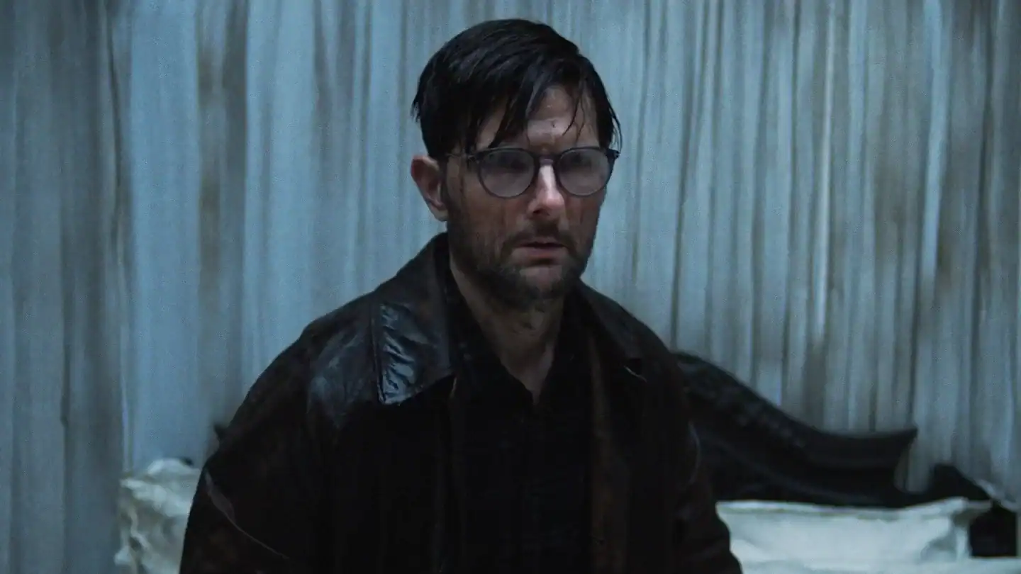A man with wet hair and glasses, wearing a dark jacket, stands indoors in front of rumpled white curtains and a bed.