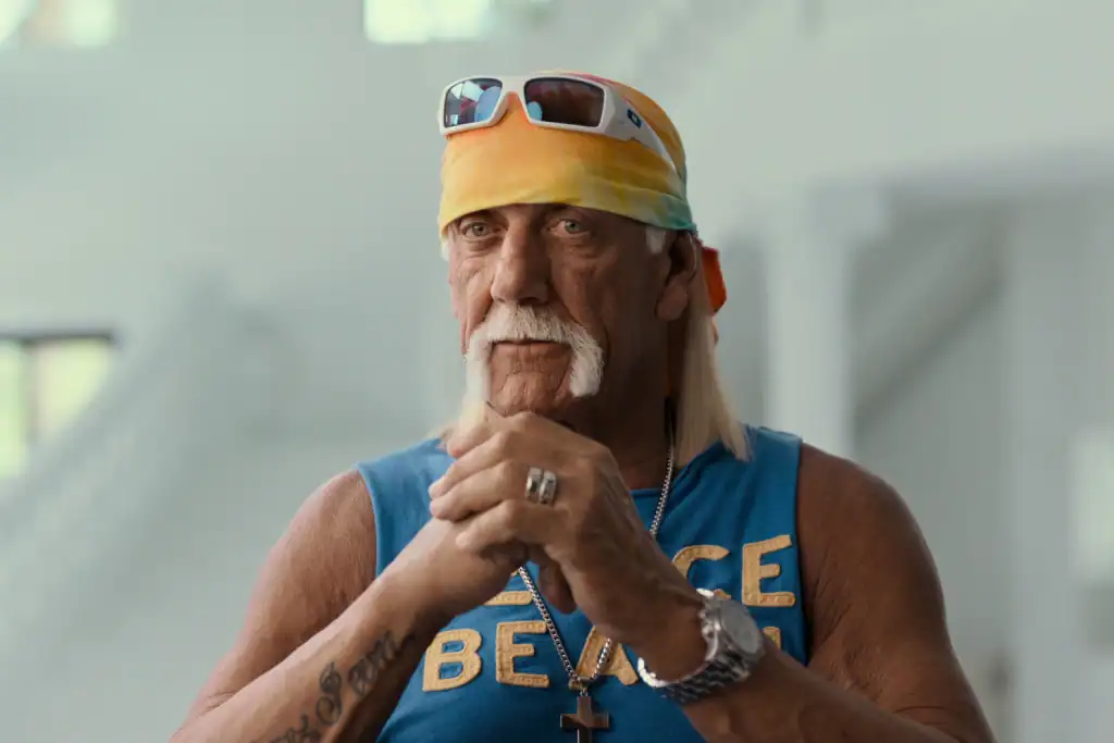 A man with a handlebar mustache, wearing a yellow bandana, sunglasses, blue sleeveless shirt, and jewelry, standing indoors with hands clasped. Hulk Hogan: Real American
