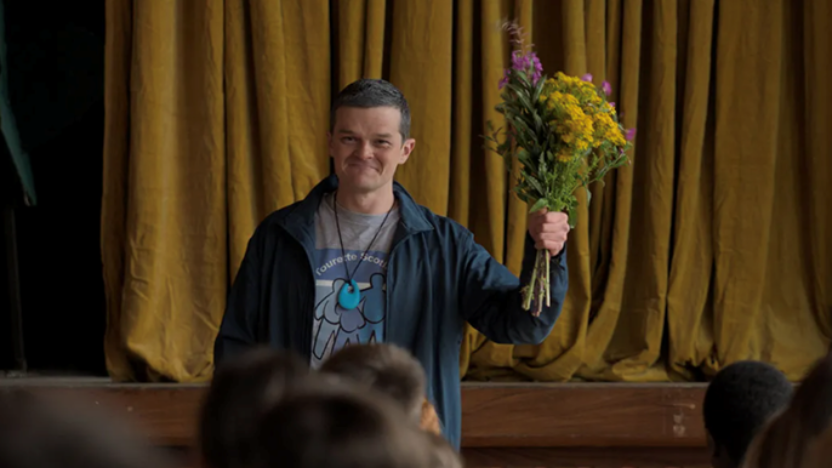A person standing in front of a yellow curtain holds up a bouquet of colorful flowers while facing an audience.