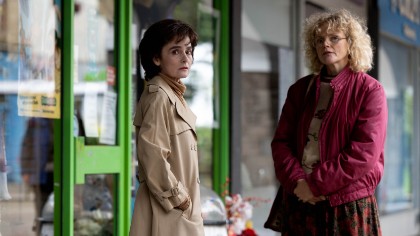 Two women stand outside a shop with green trim; one wears a beige trench coat, the other a red jacket and glasses.