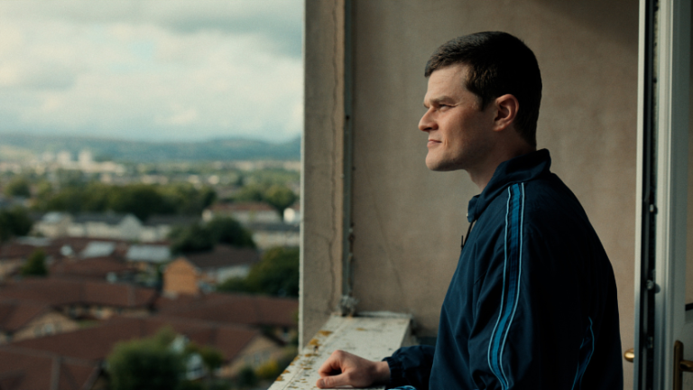 A man in a blue jacket stands on a balcony, looking out over a suburban neighborhood with houses and greenery under a cloudy sky.