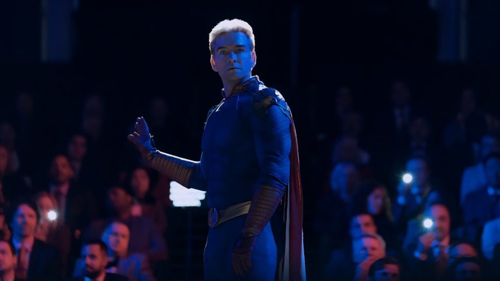 A person in a blue superhero costume with a red cape stands on stage, partially turned, with an audience watching and some people taking photos.