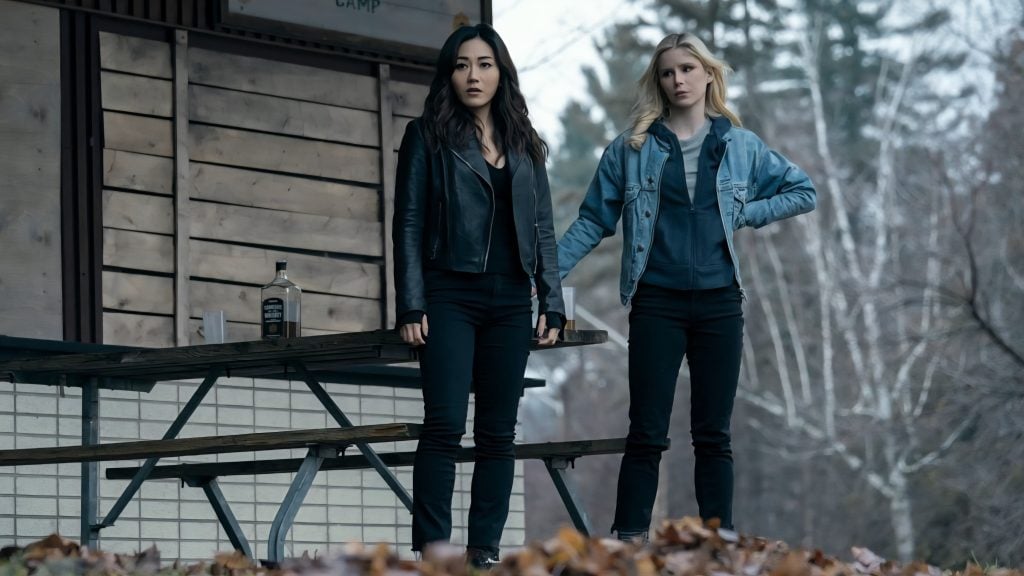 Two women stand outside near a wooden building on a cloudy day, with one resting her hand on a table holding a bottle and the ground covered in fallen leaves.