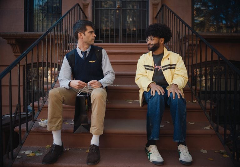 Two men sit on brownstone steps outside, facing each other. One wears a vest and tie with a bag, the other wears a yellow jacket and glasses.