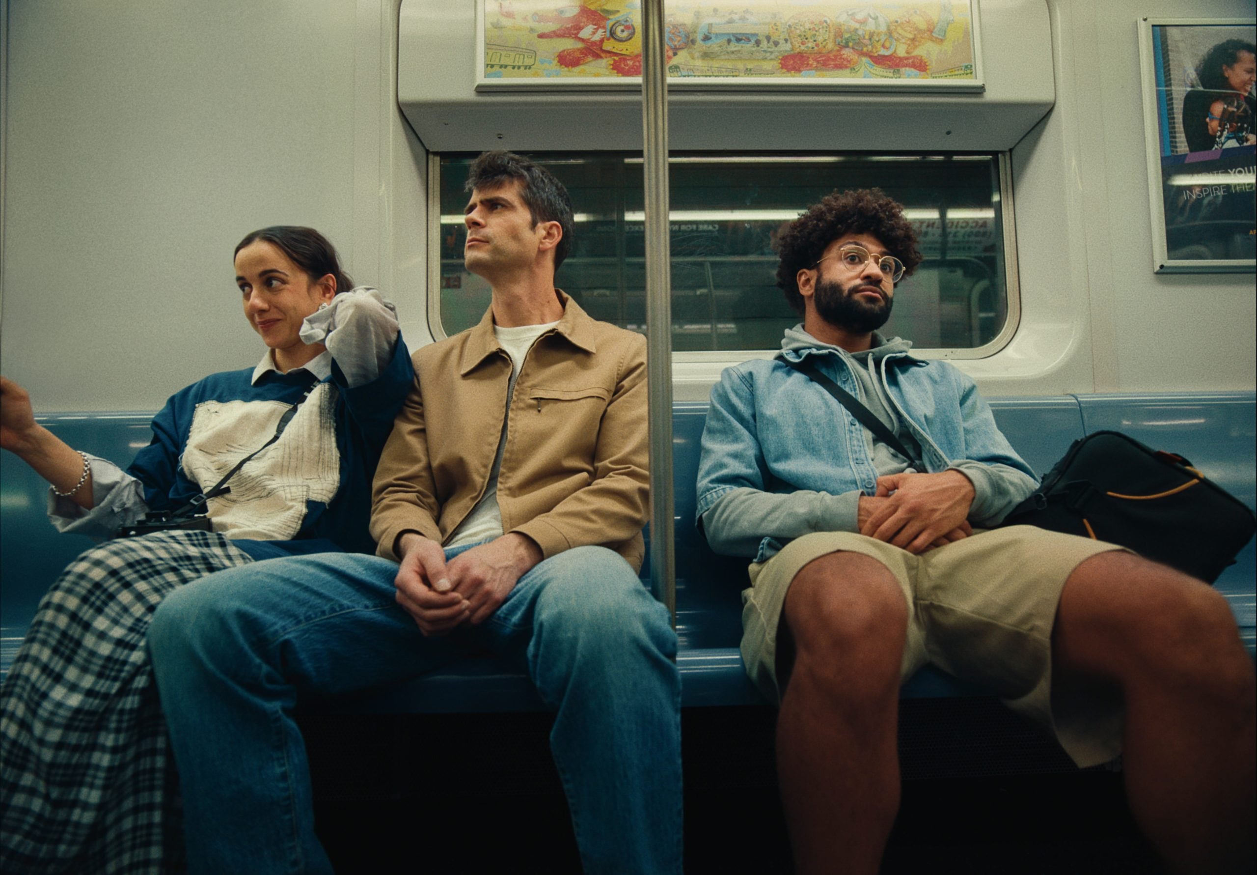 Three people sit on a subway bench; two are together on the left, the third sits apart on the right, all facing forward with neutral to pensive expressions.