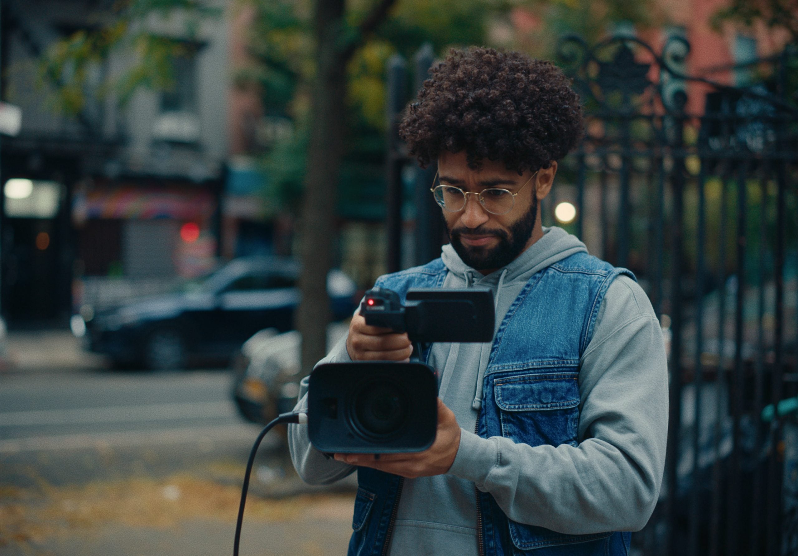 A person with curly hair and glasses operates a video camera outdoors on a city street, wearing a gray hoodie and blue denim vest.
