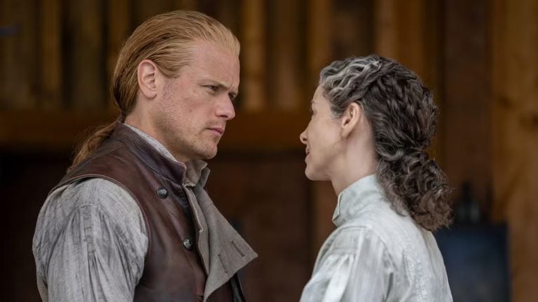 Two people dressed in period clothing stand closely facing each other, with serious and affectionate expressions, against a wooden background.