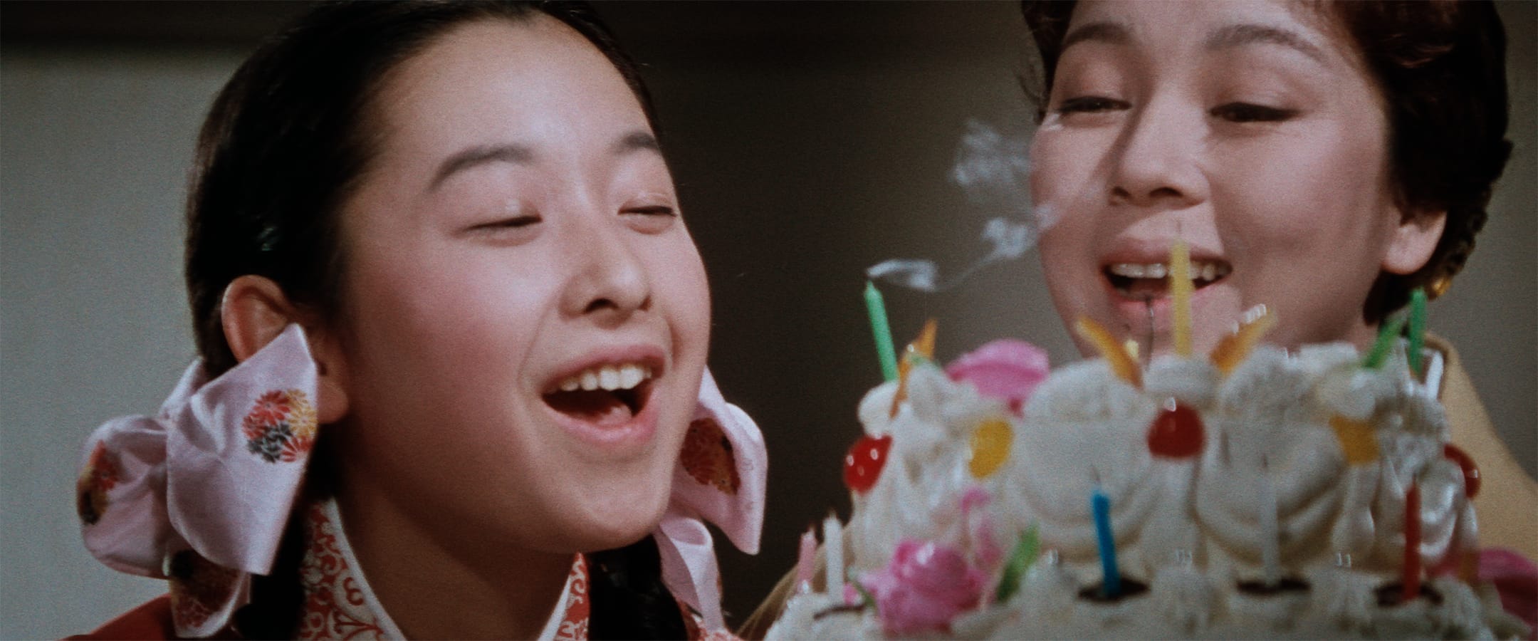 A girl with pink ribboned pigtails blows out candles on a decorated birthday cake while a woman smiles beside her.