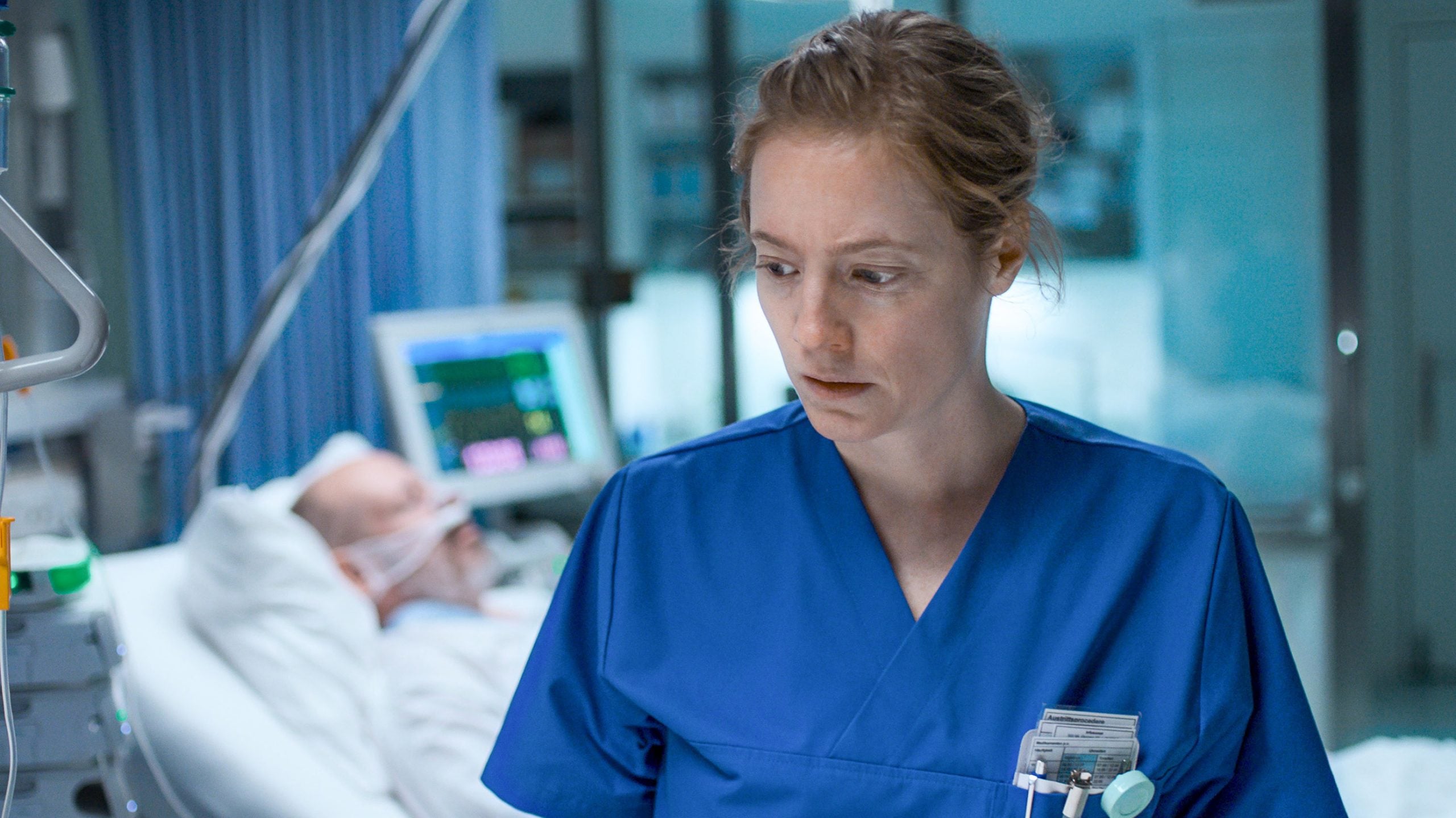 A nurse in blue scrubs stands in a hospital room, looking concerned; a patient lies in a hospital bed in the background connected to medical equipment.