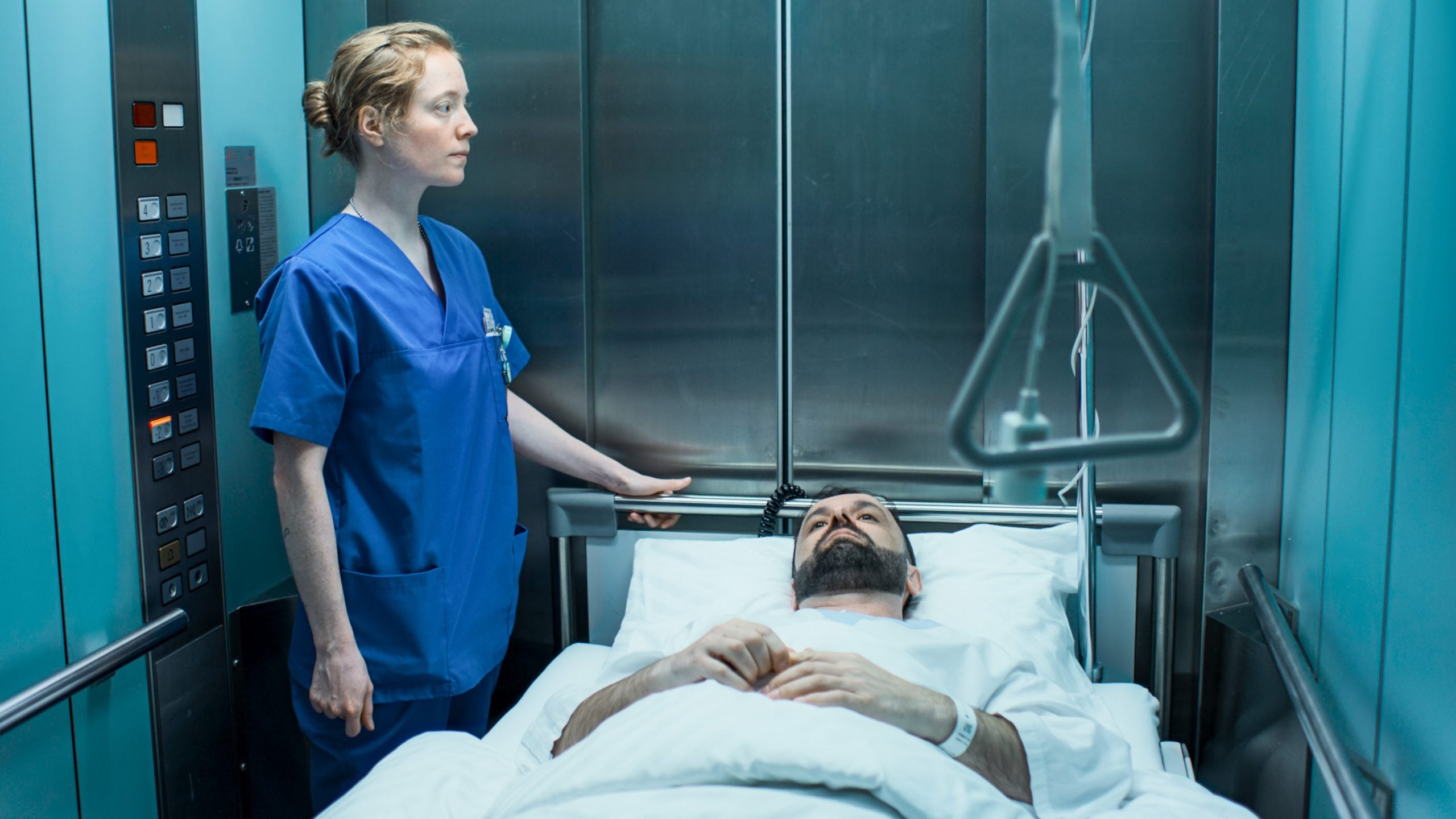 A nurse stands beside a patient lying on a hospital bed inside an elevator, with medical controls visible on the wall.
