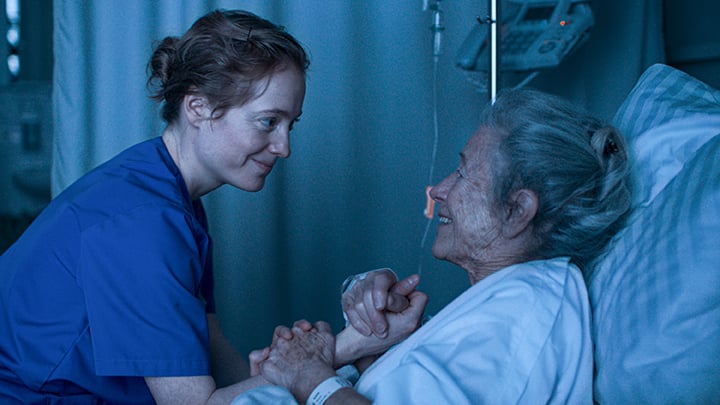 A nurse in blue scrubs holds the hand of an elderly woman lying in a hospital bed, both smiling at each other in a dimly lit hospital room.