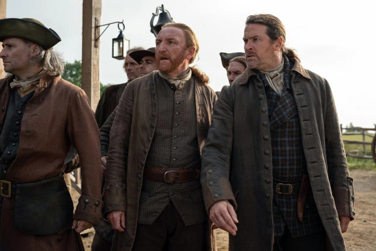 Several men in colonial-era clothing stand together outdoors, looking in the same direction with serious expressions.
