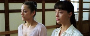Two women stand side by side indoors, facing forward. Both wear light-colored tops and have their hair tied back. Wooden framed doors and windows are visible in the background.