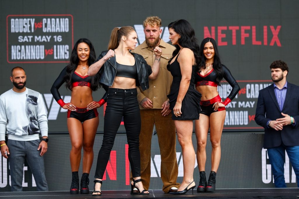 Two female fighters face off on stage with fists raised, flanked by two ring girls and three men, in front of a backdrop with Netflix logos and fight promotion details.