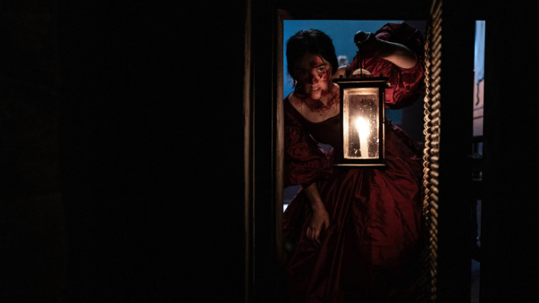 A woman in a dark red dress holds a lit lantern while standing in a dimly lit doorway, partially illuminated by the lantern’s glow.