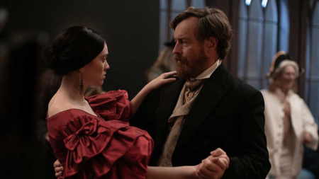 A woman in a red dress and a man in a black suit and beige cravat are dancing together in a dimly lit room with arched windows in the background.