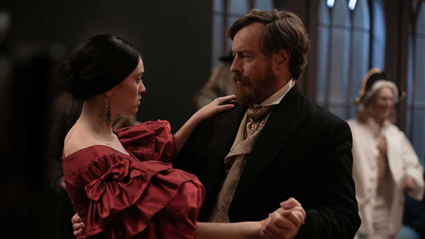 A woman in a red dress and a man in a black suit and beige cravat are dancing together in a dimly lit room with arched windows in the background.