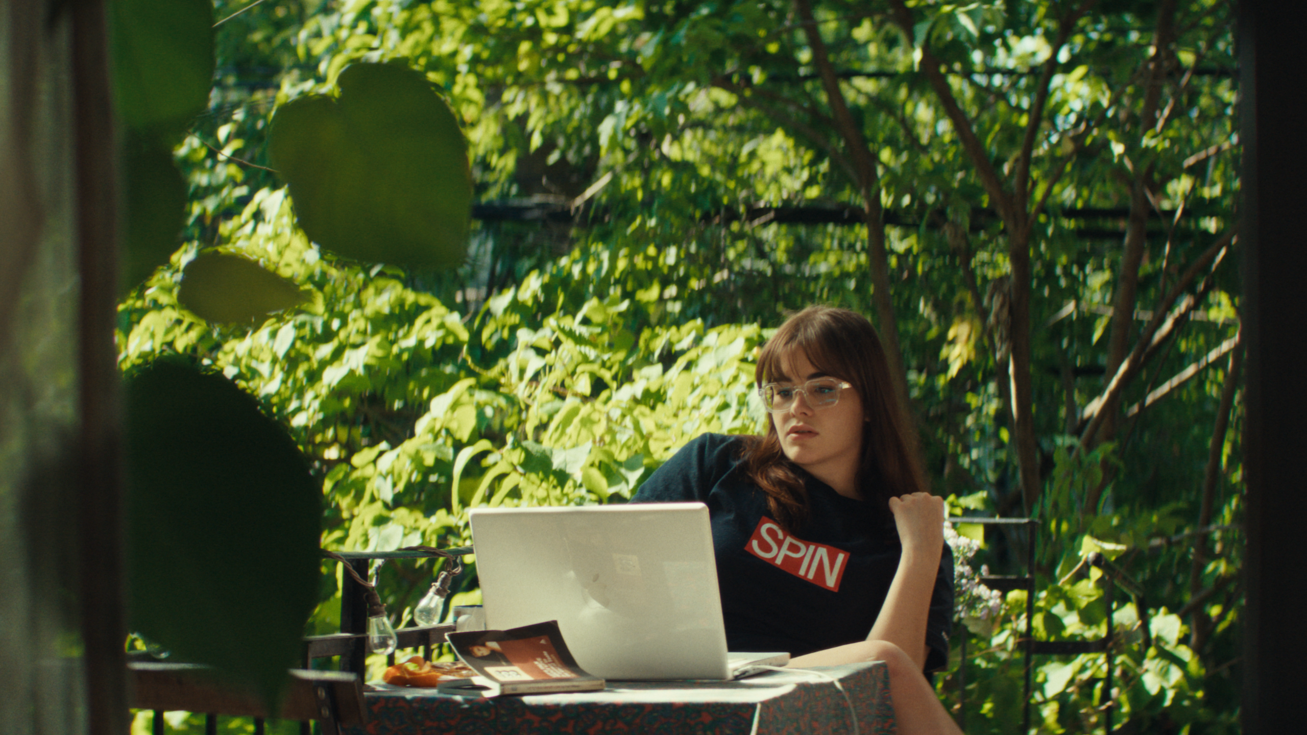 A person with glasses sits at an outdoor table with a laptop, book, and fruit, surrounded by green foliage.