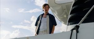 ‘Mermaid’ Blu-Ray Review – A Wonderfully Scuzzy, Sun-Soaked Creature Feature A man in casual clothing stands on the deck of a white boat, looking down with a serious expression against a cloudy sky.