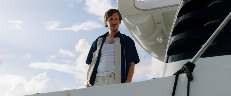 ‘Mermaid’ Blu-Ray Review – A Wonderfully Scuzzy, Sun-Soaked Creature Feature A man in casual clothing stands on the deck of a white boat, looking down with a serious expression against a cloudy sky.