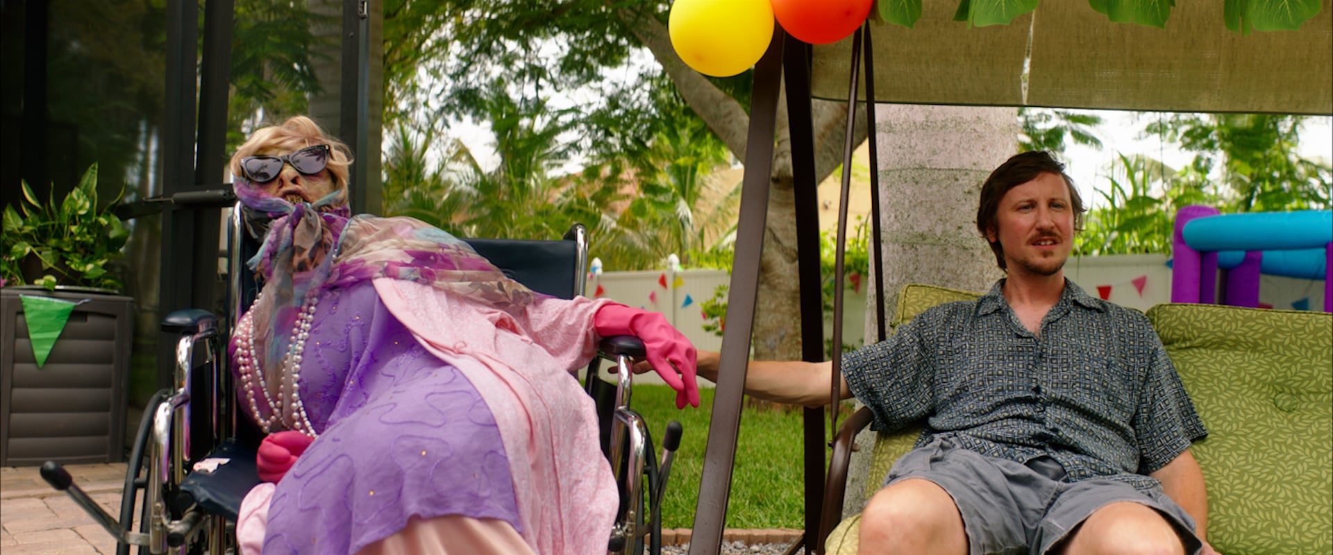An older woman in a wheelchair and a man sit outside under balloons, surrounded by greenery and party decorations.
