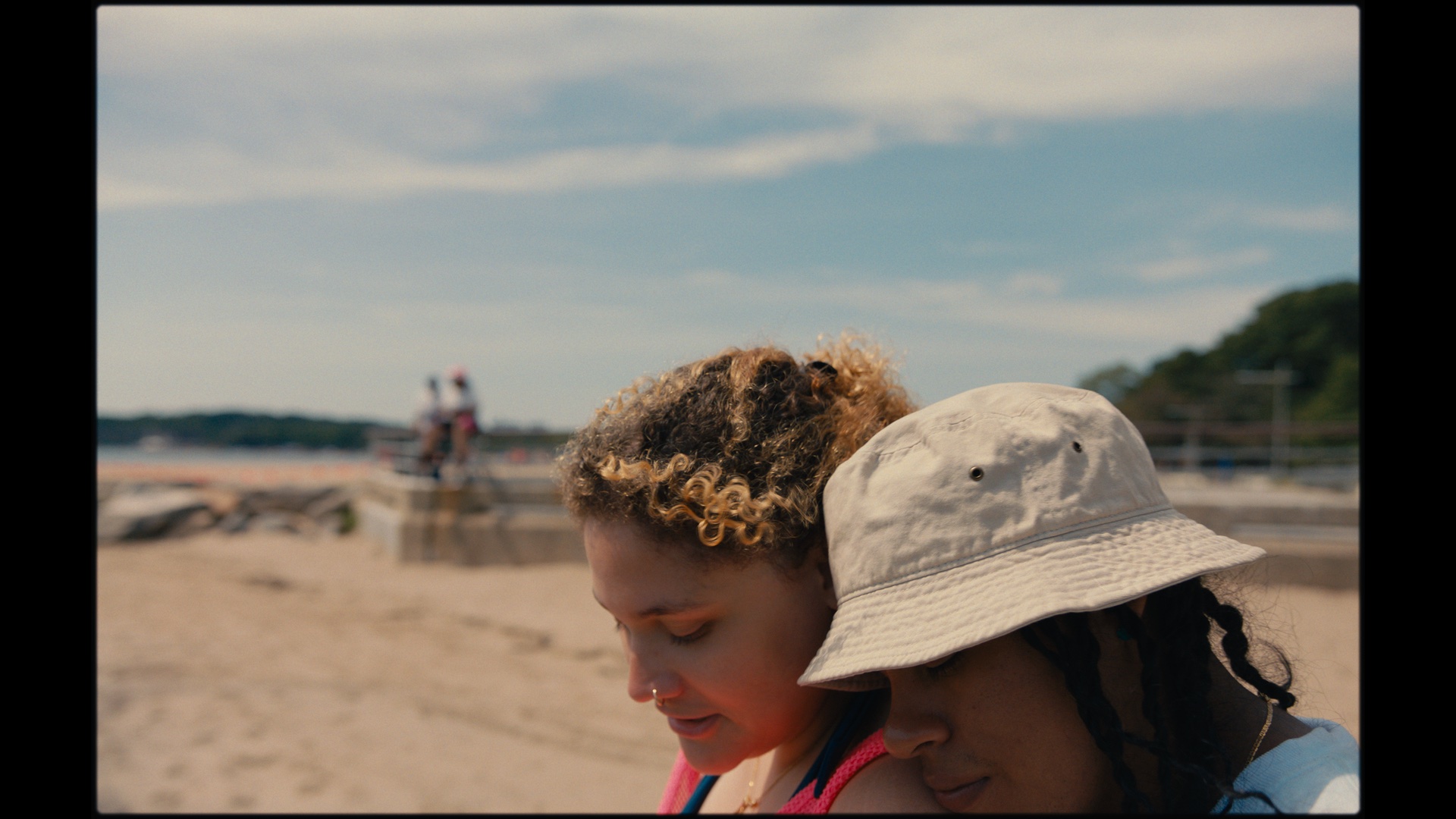 Two people with curly hair stand close together on a sandy beach, one wearing a light bucket hat. The background shows a beach scene with people and a blue sky.