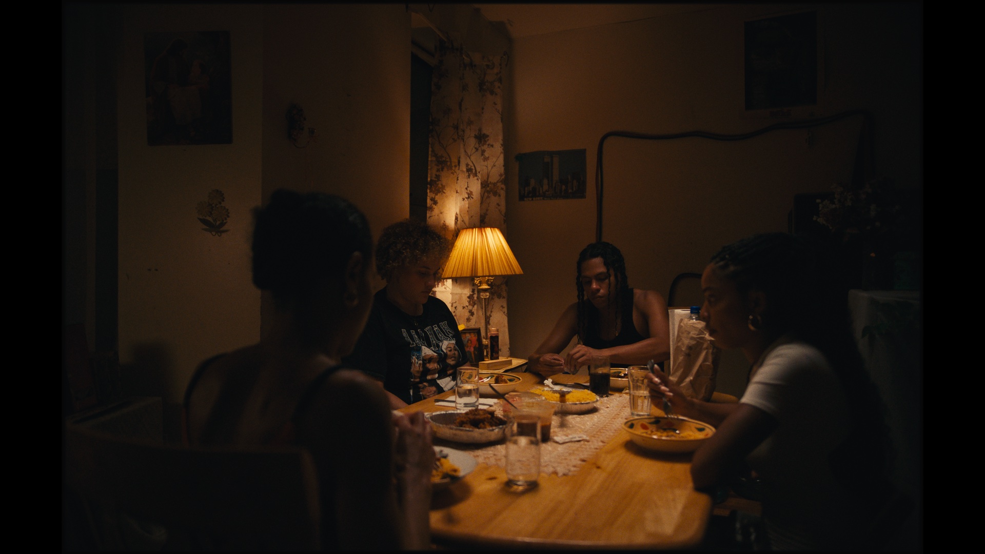 Four people sit around a wooden dining table eating a meal together in a dimly lit room with a lamp and floral wallpaper.