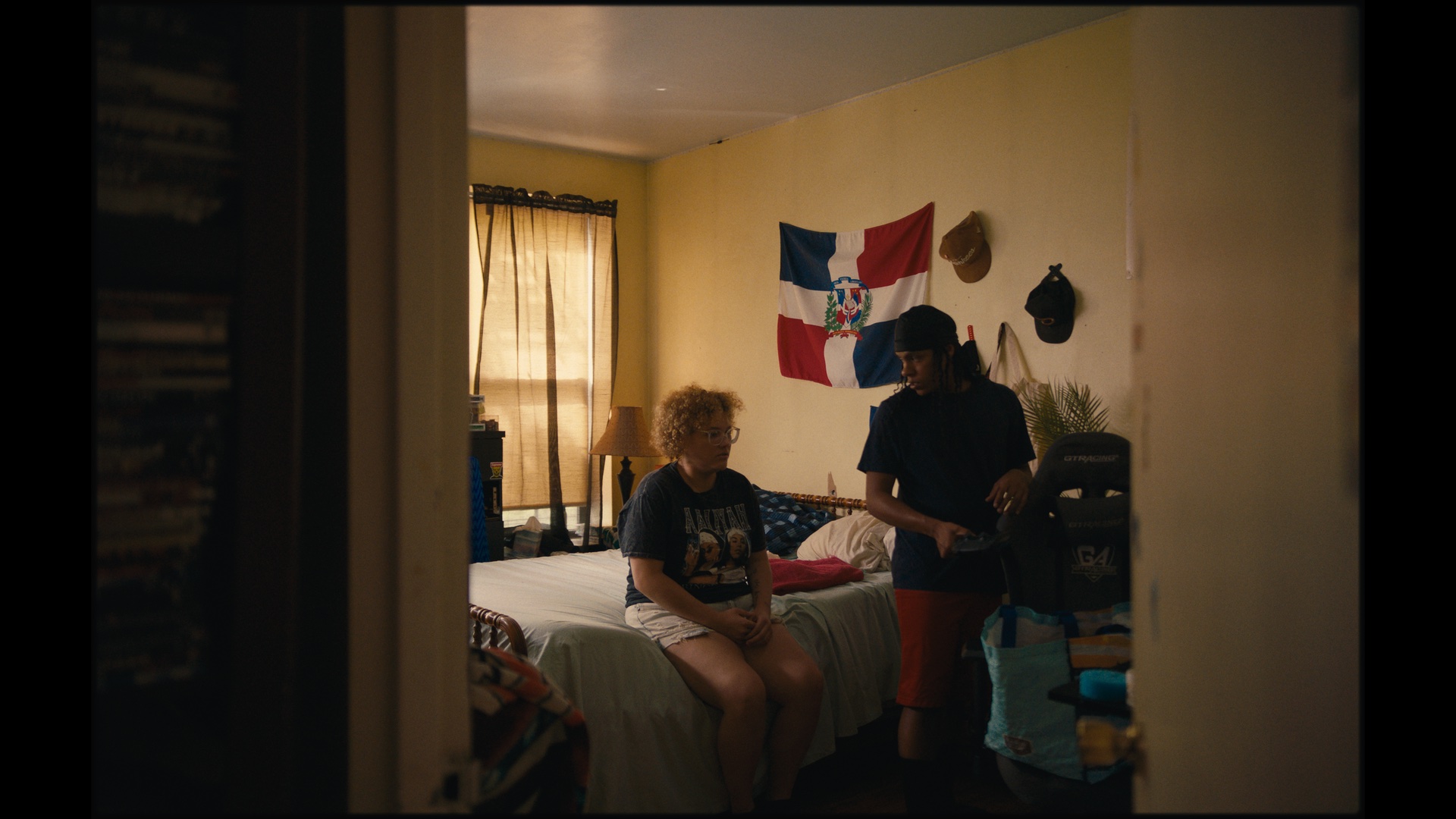 Two people are in a bedroom with yellow walls, a bed, a Dominican Republic flag on the wall, hats, and sports gear. One person sits on the bed while the other stands beside it.