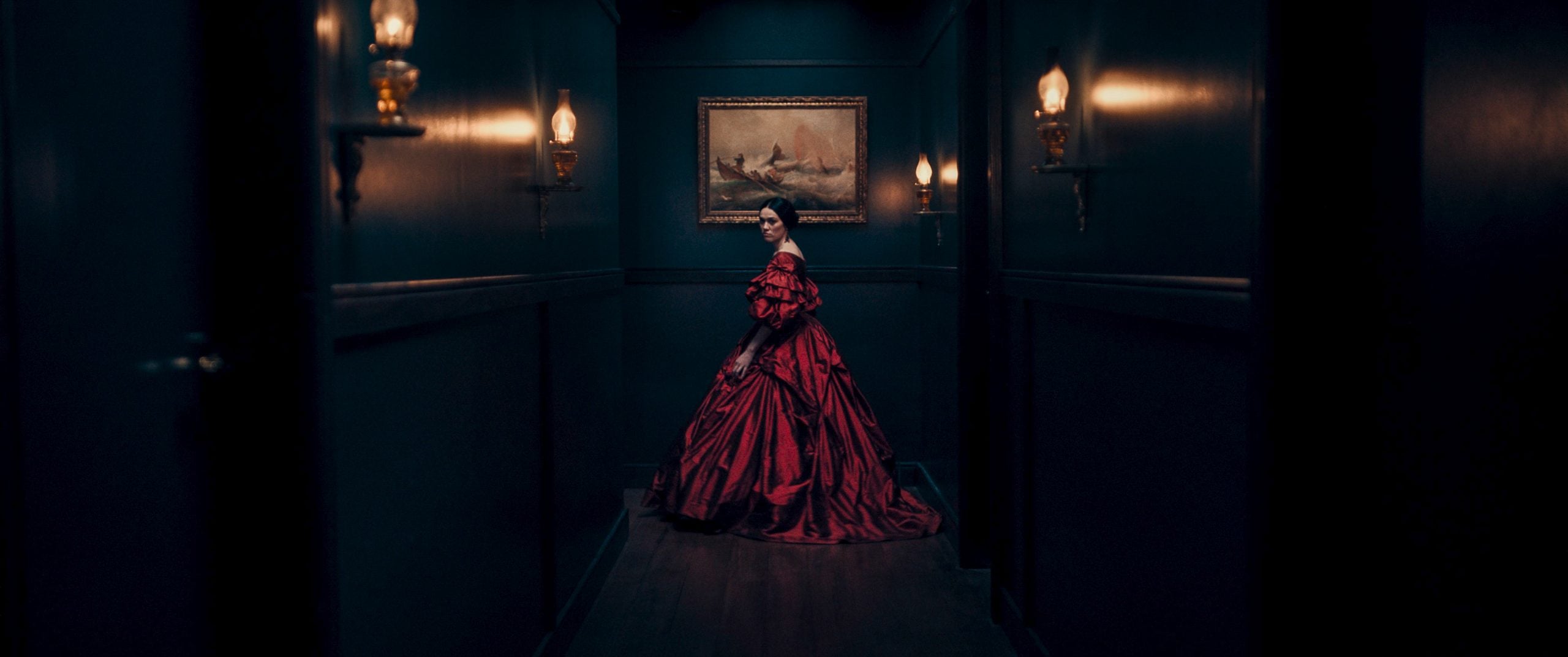 A woman in an elaborate red dress stands in a dimly lit hallway, facing away from the camera, with a framed painting on the wall in front of her.