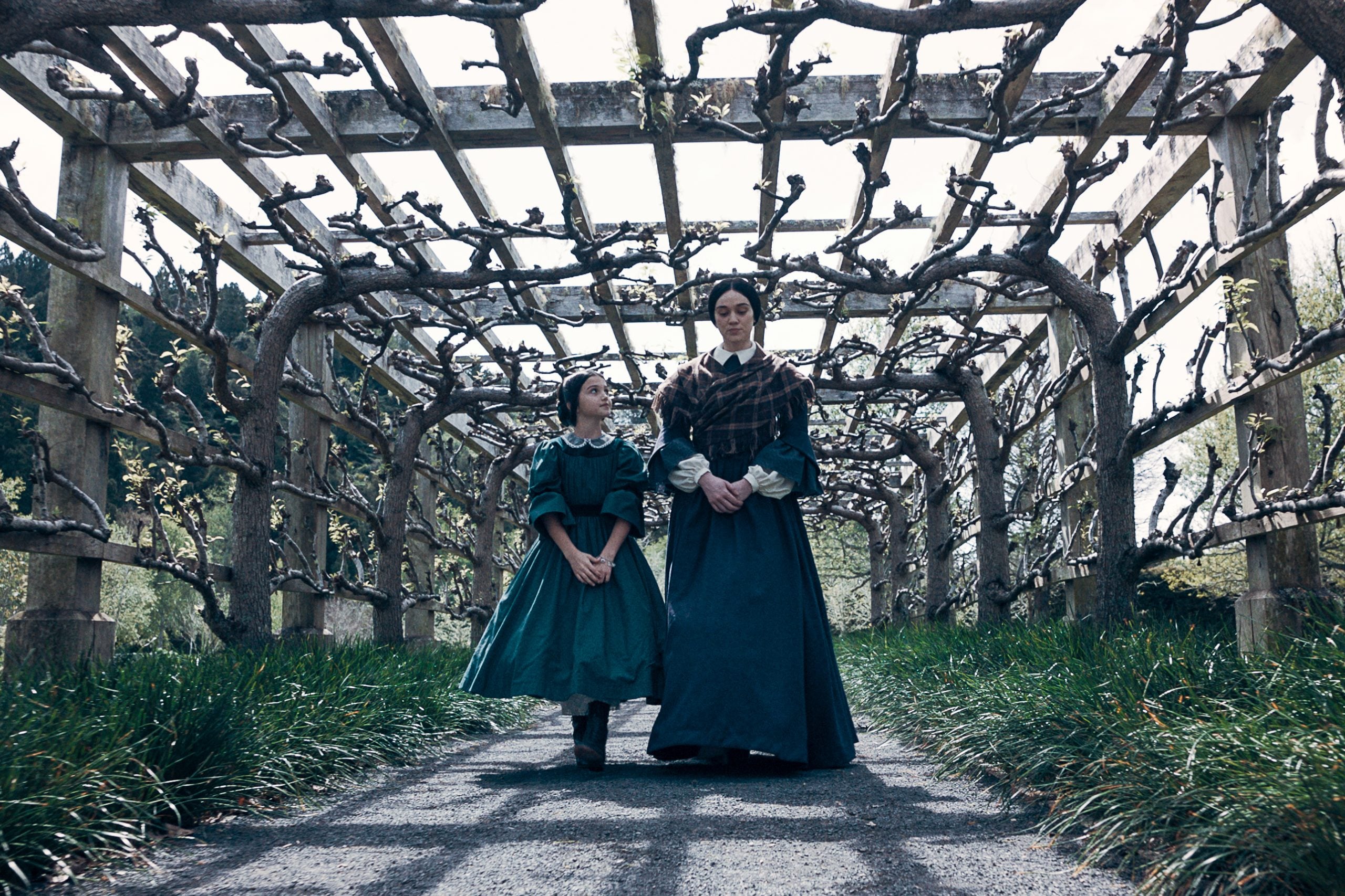Two women in period clothing walk under a wooden trellis with bare, pruned vines on a paved path, surrounded by greenery.