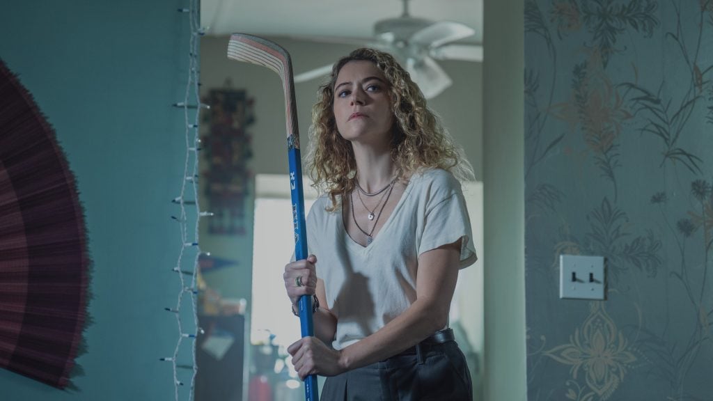 A woman with curly hair, wearing a white shirt and dark pants, stands indoors holding a blue hockey stick, looking directly ahead.