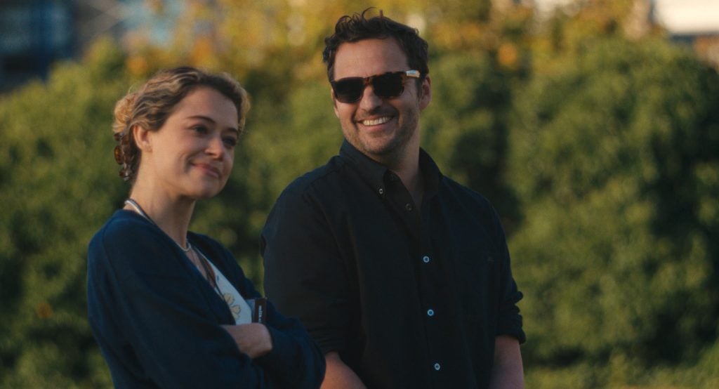 Two people standing outdoors, one smiling and wearing sunglasses, the other with arms crossed, both appearing relaxed in front of a blurred green background. Maximum Pleasure Guaranteed
