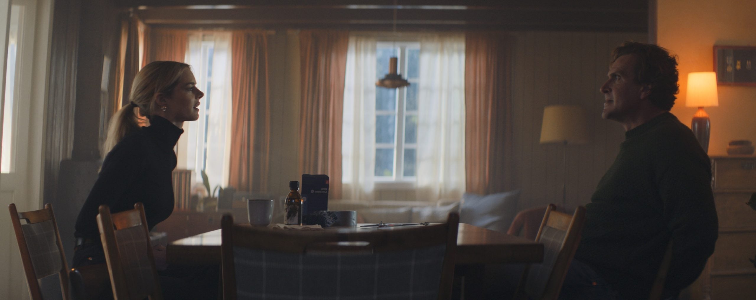 Two people sit on opposite sides of a dining table in a dimly lit room, appearing to be in a serious conversation.
