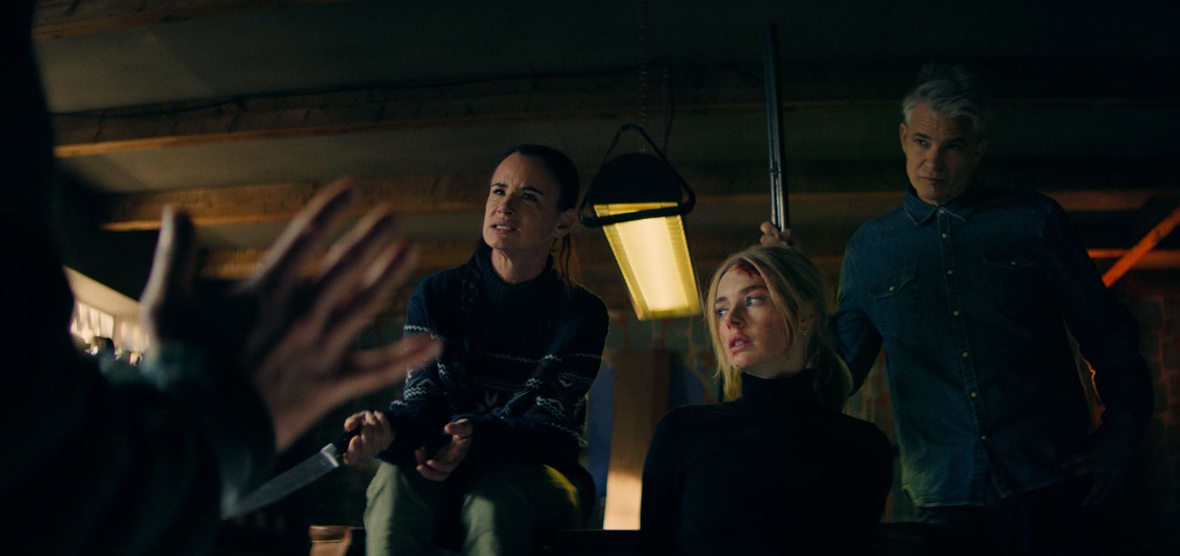 Three people indoors, two sitting and one standing, with serious expressions; one person holds a shotgun and another faces a figure with raised hands in the foreground.