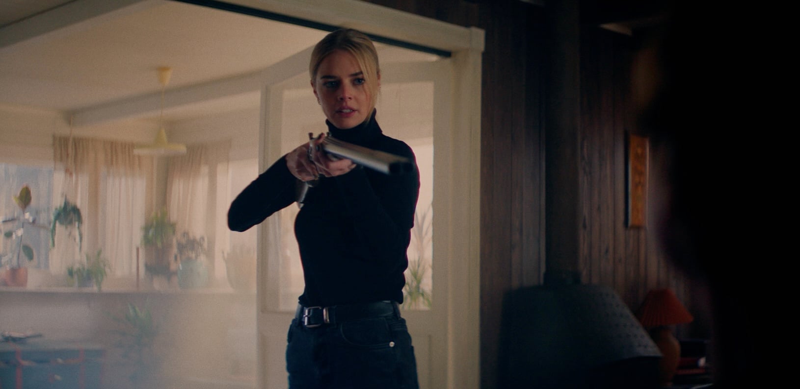 A woman in a black turtleneck stands indoors aiming a shotgun, with a serious expression on her face.