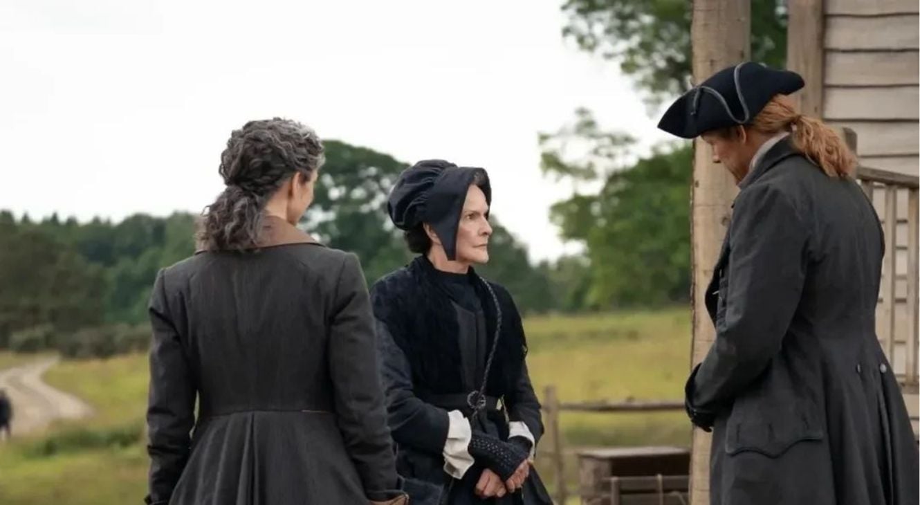 Three people in period clothing stand outside near a wooden building, engaged in conversation, with trees and a field in the background.